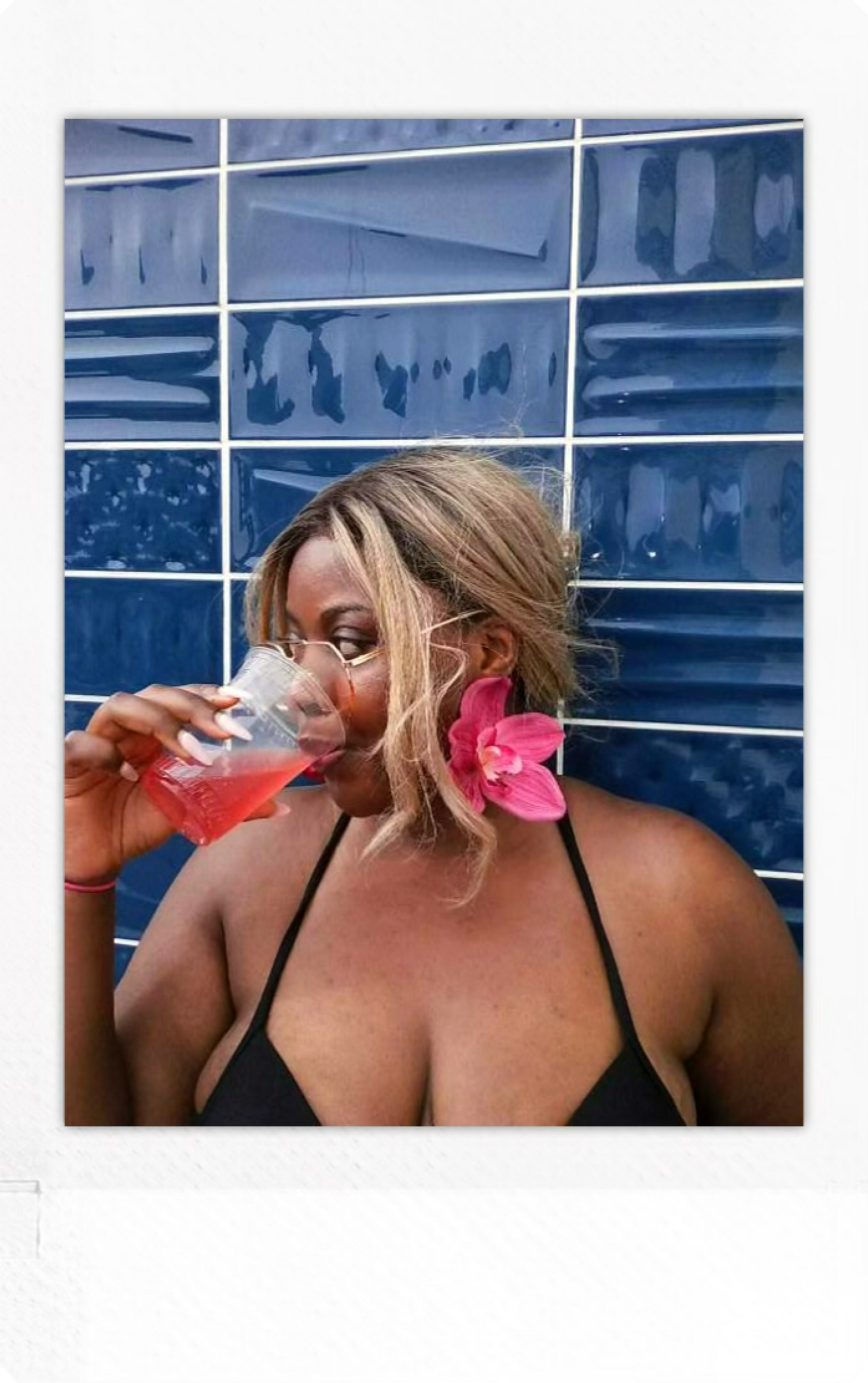 Woman drinking from a plastic cup with pink liquid against a blue tiled wall.